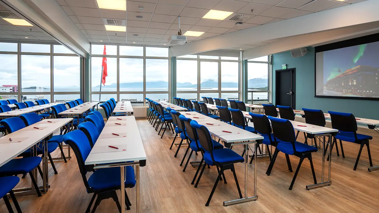 Large and bright meeting room at Thon Hotel Harstad. Tables and chairs in a classroom layout. Large windows in the background.