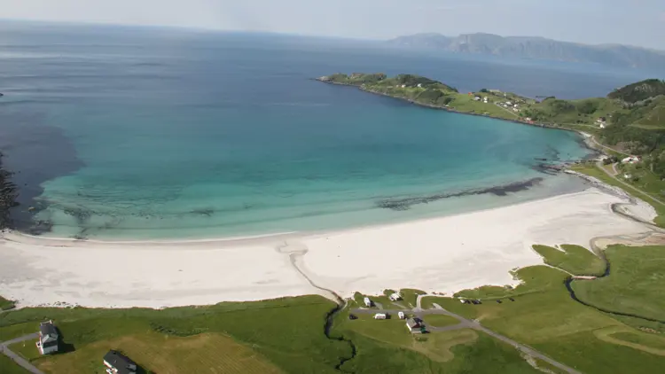 Aerial view of bay with white sandy beach and crystal blue water surrounded by green landscape.