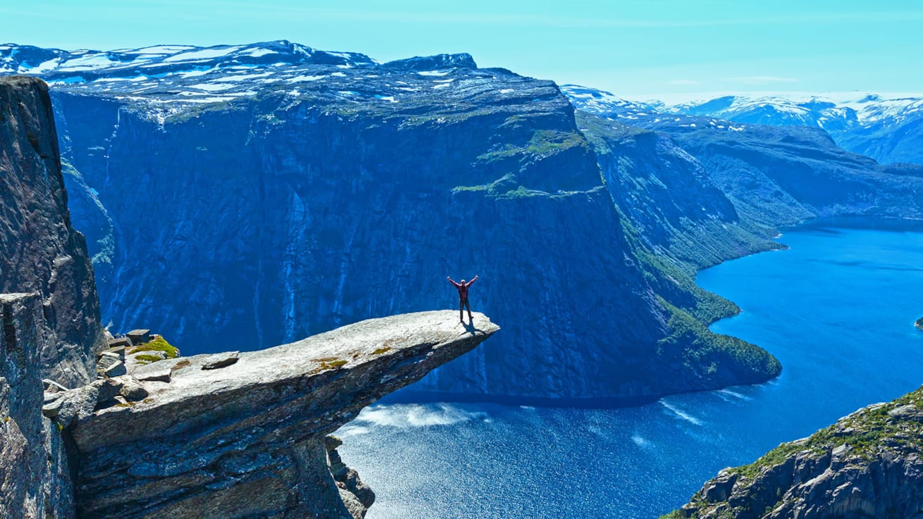 En person st&aring;r ytterst p&aring; Trolltunga i str&aring;lende sol